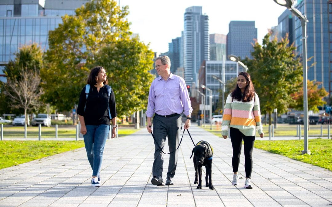 A group of three people walking outdoors amidst a city background. A man in the middle is holding a dog on a leash.